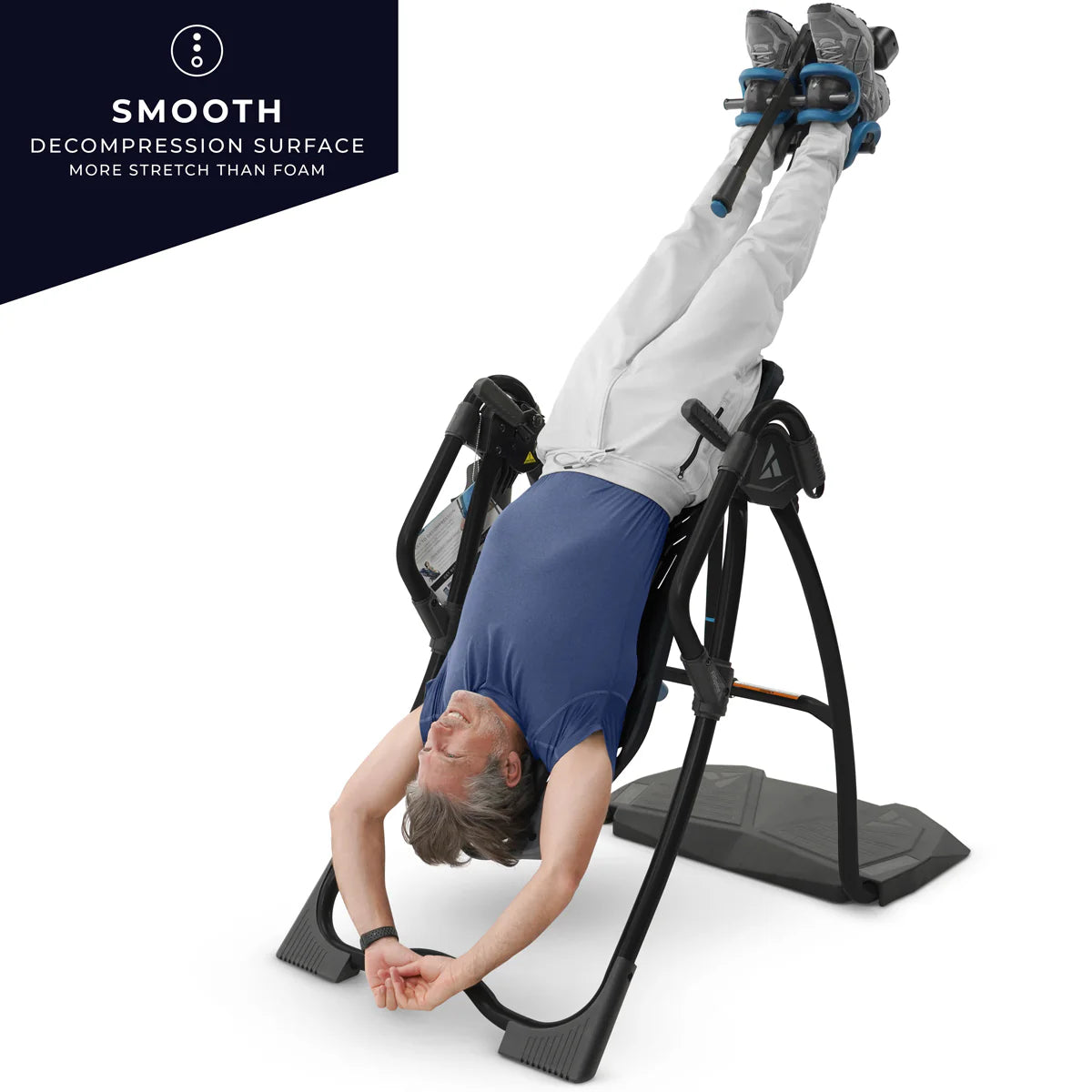 Middle aged male demonstration the Teeter Fit Spine Inversion Table. They are suspended in the device with their feet secured at the top and their head towards the bottom in an upside down position. The male in dress in casual athletic clothes. White jogging bottoms, dark blue trainers and grey trainers. Text to the top left of the screen reads SMOOTH decompression surface more stretch than foam.